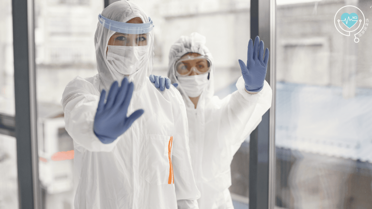 Photo of two healthcare workers wearing full protective suits, face masks, face shields, and gloves, raising their hands as if signaling to stop or maintaining safety precautions.